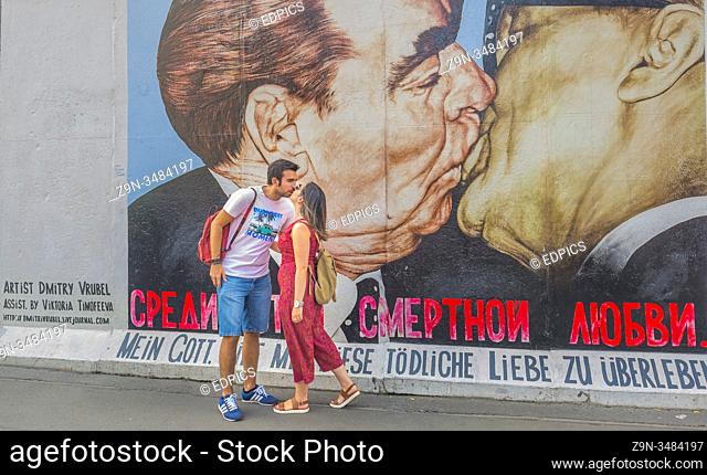 young couple kissing in front of mural painting at east side gallery at east side gallery, friedrichshain-kreuzberg, berlin, germany