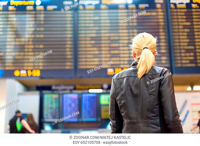 Casually dressed young stylish female traveller checking a departures board at the airport terminal hall in front of check in couters