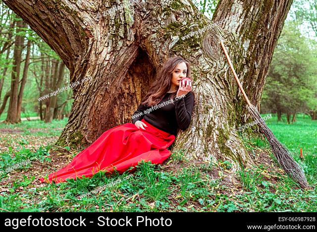 mystical witch woman in red dress sitting under a old tree in the forest