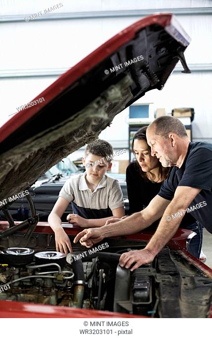A senior Caucasian male car mechanic talks to a Caucasian mother and her son about a engine repair issue in a classic car repair shop