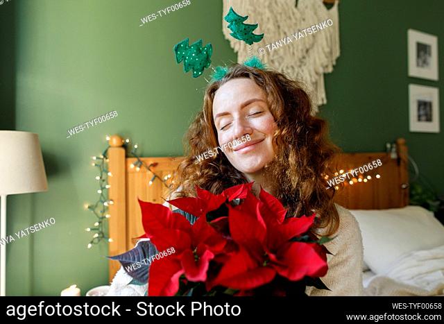 Smiling woman with eyes closed holding Poinsettia plant at home