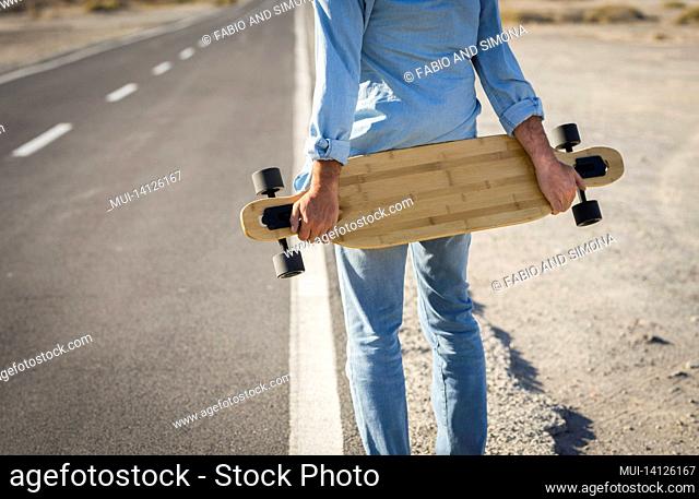 hands of man holding skateboard on road. rear view of man wearing denim clothes holding skateboard on rural road. sportsman holding skateboard and waiting on...