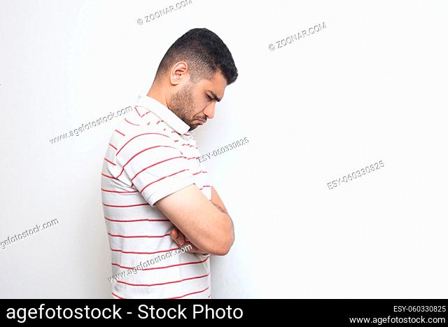 Profile side view portrait of sad alone o depressed bearded young man in striped t-shirt standing, holding his head down and feeling bad or thinking