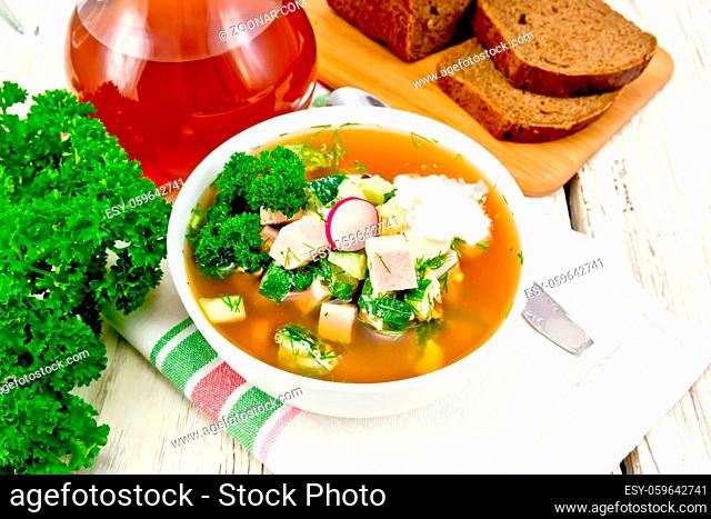 Cold soup okroshka from sausage, potatoes, eggs, radish, cucumber, greens and kvass in a white bowl on napkin, bread and jug with drink on the background of...