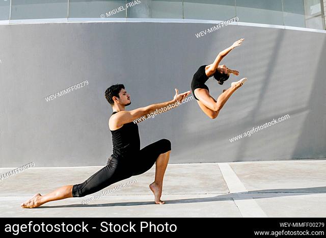 Female gymnast doing acrobat while male dancer practicing stag leap by gray wall