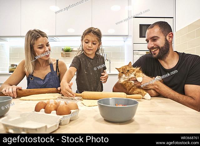 Mother and daughter kneading dough with rolling pin while father holding cat on kitchen's table
