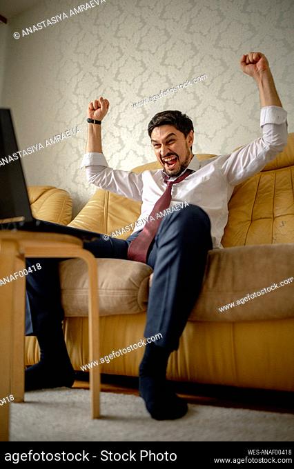 Cheerful businessman gesturing fists sitting on sofa at home