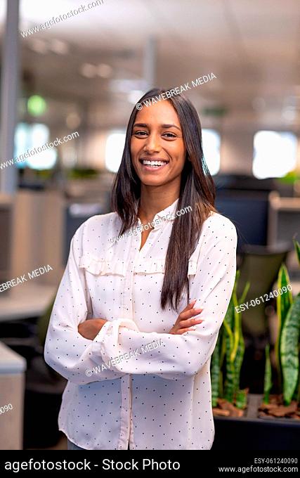 Portrait of beautiful happy biracial businesswoman standing with arms crossed at modern workplace