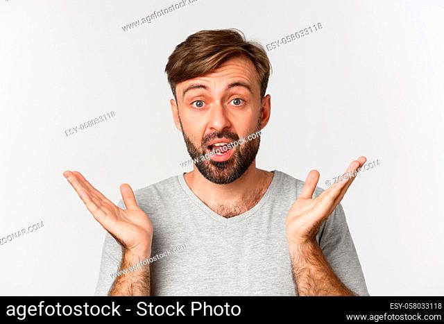 Close-up of surprised handsome man with beard, spread hands sideways and look in awe, standing over white background