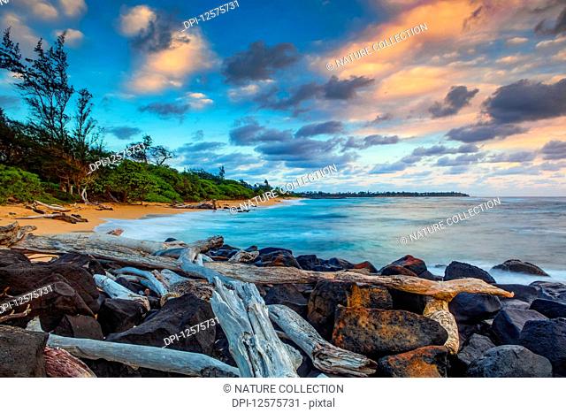 Sunrise over Lydgate beach and ocean with rocks and driftwood in the foreground and the coastline in the distance; Kapaa, Kauai, Hawaii