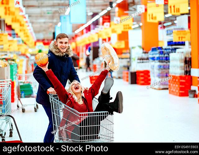 Young handsome guy rides a girl in a supermarket in a trolley. Couple have fun. Girl holds an orange and bread