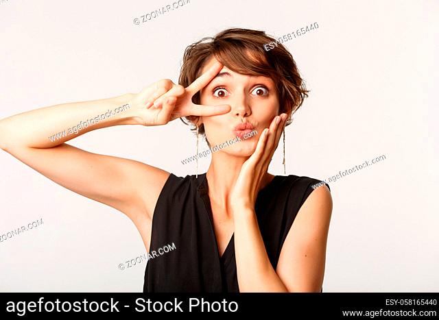 Close-up of silly beautiful woman pouting and showing peace gesture over eye, standing against white background