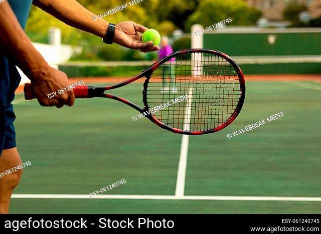 Midsection of biracial senior man holding racket serving tennis ball while playing at tennis court