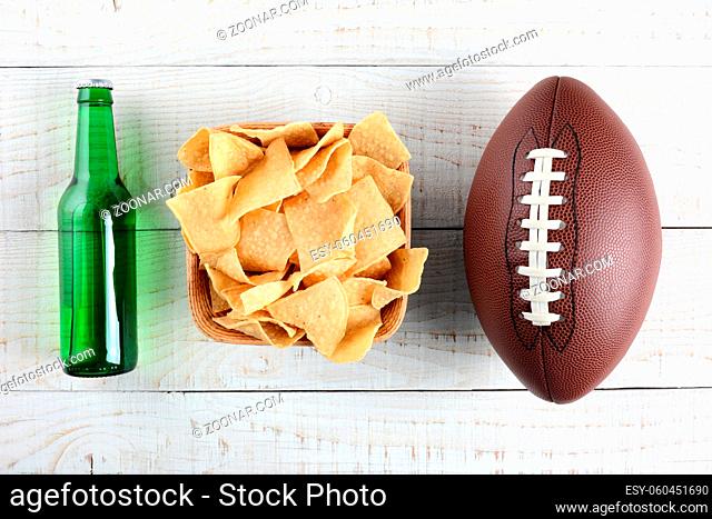 Beer bottle, bowl of chips and an American style football on a rustic whitewashed wood surface. Horizontal format. The bottle is without label