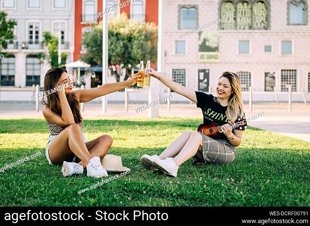 Cheerful female friends toasting drinks while sitting on grassy land