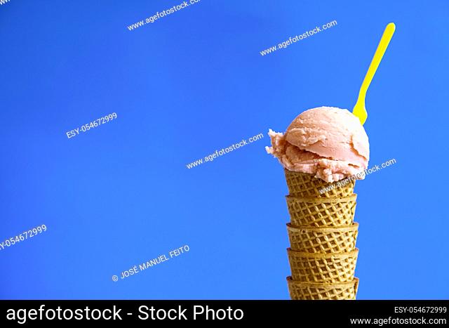 strawberry ice cream ball on waffle cornet with yellow plastic teaspoon on blue background. Copy space