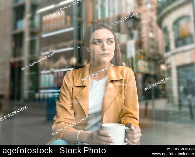 Thoughtful young woman sitting at cafe seen through glass window