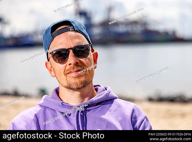 PRODUCTION - 31 May 2022, Hamburg: The musician Jan Delay sits on a cafe stool on the Elbe beach in Ovelgönne in front of the industrial facilities of the port...