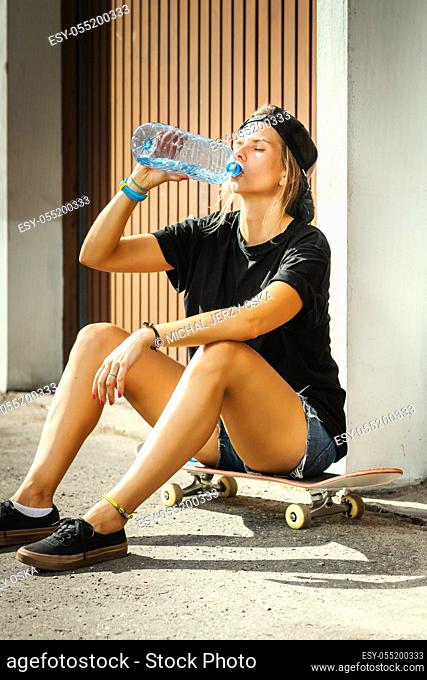girl is sitting on a skateboard and is drinking water on a summer day