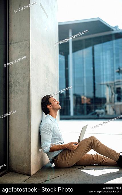 Businessman with laptop looking up sitting by by wall outside building