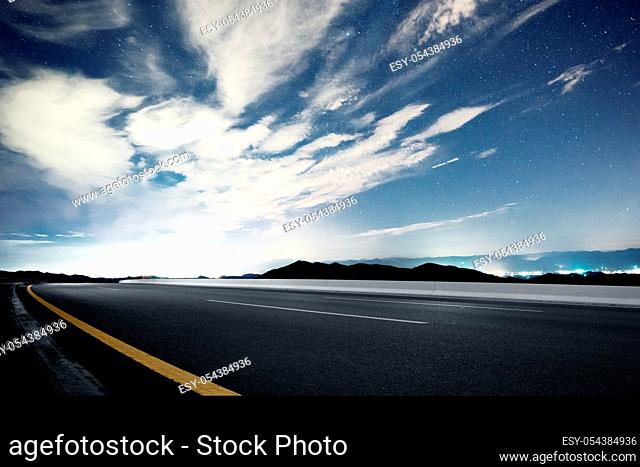 empty asphalt road passed through illuminated modern city surrounded by green hill in blue cloud star sky