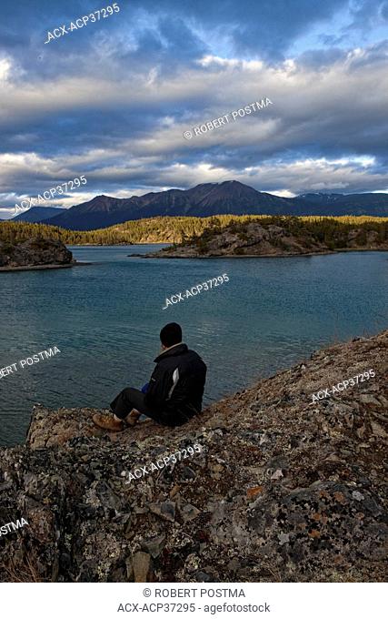 Man sitting on rocks watching the sunset light hitting an island on Atlin Lake in Atlin Provincial Park, Atlin, British Columbia, Canada