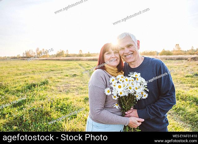 Happy senior couple holding chamomile flowers at field on sunny day