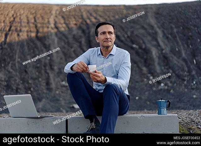 Mature businessman sitting on stairs on a disused mine tip next to laptop having a coffee break