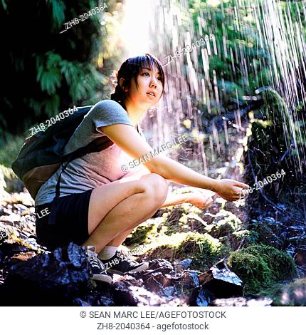 A girl with a backpack and hiking shoes holding her hands out under a waterfall off the Multnomah-Wahkeena Loop Trail in Oregon