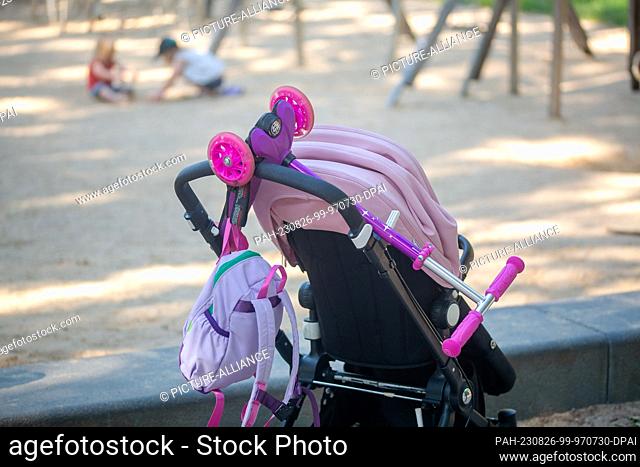 PRODUCTION - 08 July 2023, Berlin: A stroller with a scooter and a child's backpack hanging from it stands next to a playground while two toddlers play in the...