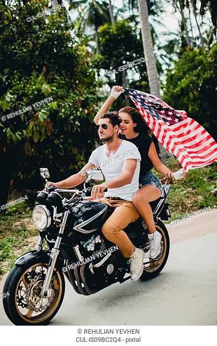 Young couple holding up American flag while riding motorcycle on rural road, Krabi, Thailand