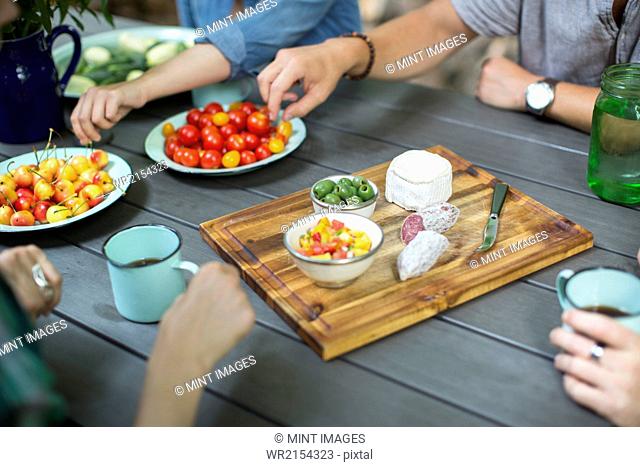 A group of people gathered around a table with plates of fresh fruits and vegetables, and a round cheese and salami on a chopping board