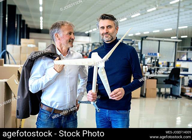 Smiling businessman holding wind turbine model standing by colleague at warehouse