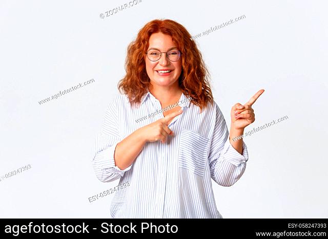 Portrait of pleasant smiling middle-aged woman with red hair, wearing glasses and blouse showing advertisement, client of company recommend product or service