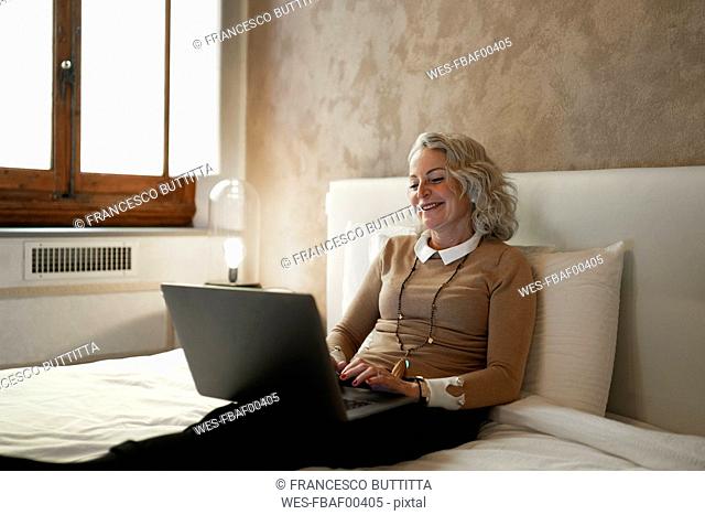 Portrait of content mature businesswoman sitting on bed working on laptop
