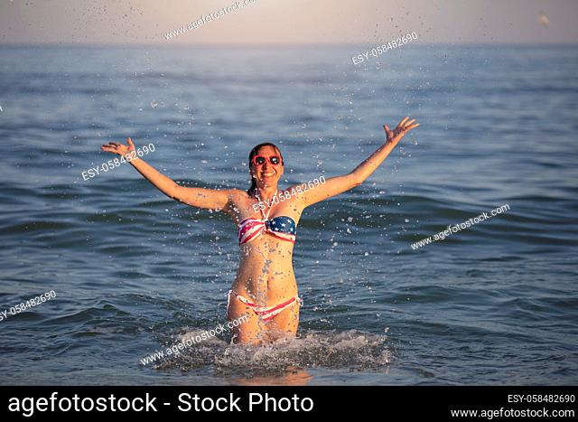 Happy middle aged woman splashing sea water in swimsuit with American flag print