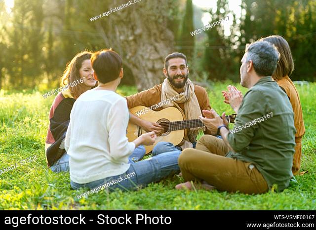 Group of friends playing music with the guitar sitting on the grass in the field