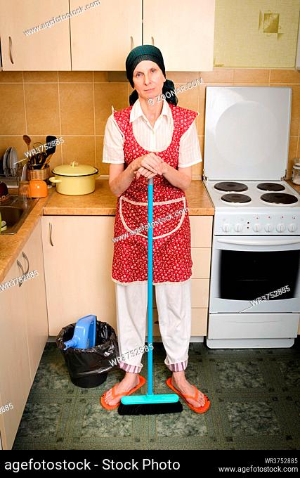 A depressed adult woman, a housewife or a maid, wearing a red apron and a green scarf on her head is resting after she has swept the kitchen with a broom