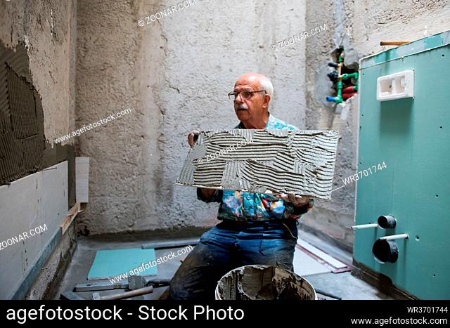 experienced senior construction worker installing big ceramic tiles on interior concrete walls