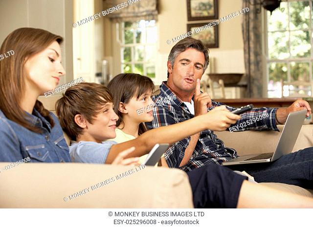 Children Watching TV Whilst Parents Use Laptop And Tablet Computer At Home