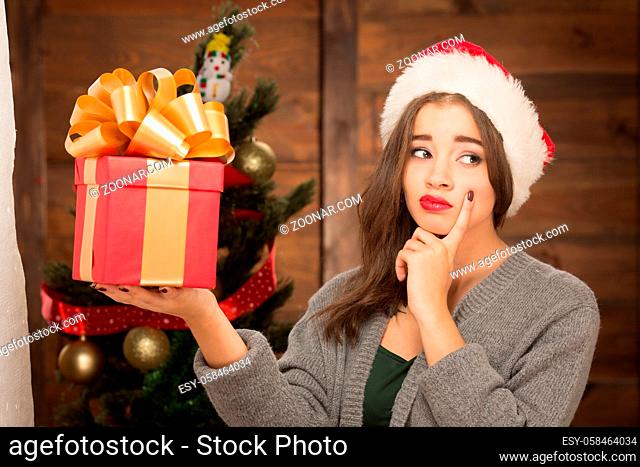 Serious girl holding a present in front of New Year tree and thinking about possible gifts in it. Pretty lady looking away and touching her cheek