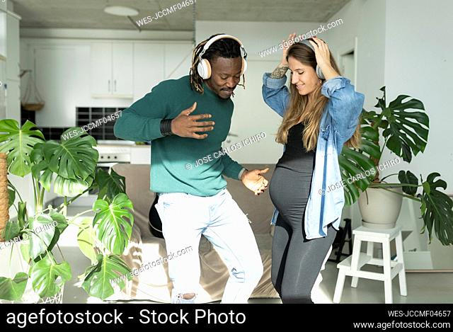 Man and pregnant woman wearing headphones dancing together in living room