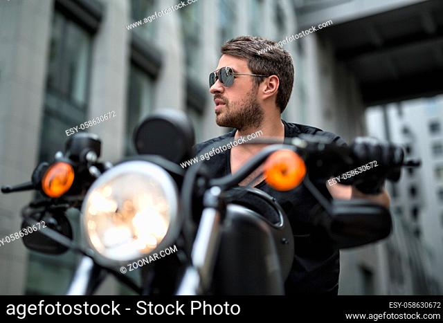 Charismatic man sits on the black motorbike on the skyscraper background. Headlamp switched on. He wears a black T-shirt, black gloves and sunglasses
