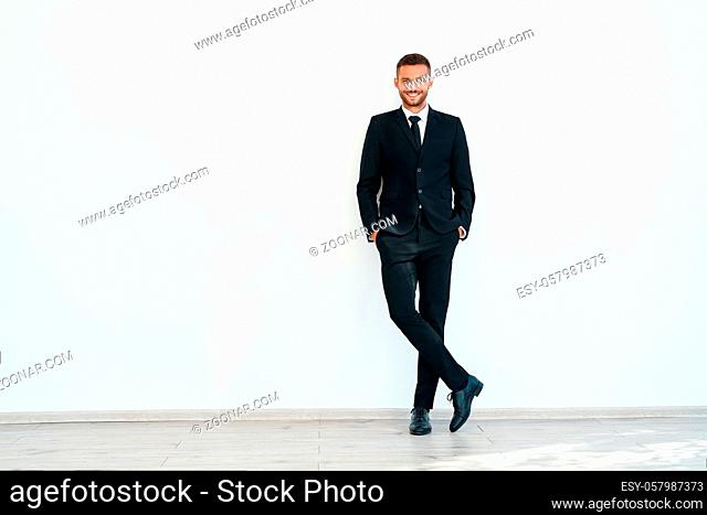 Full length portrait of smiling confident businessman in elegant suite posing on white wall background with copy space