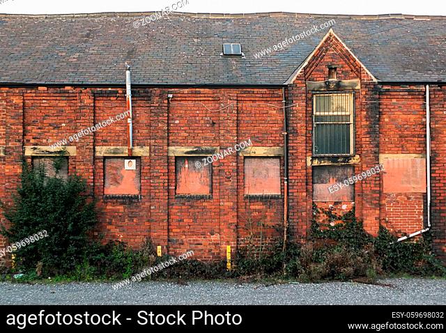 An old abandoned brick factory building with boarded up windows and crumbling decaying walls overgrown with weeds