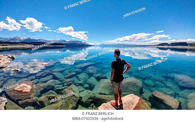 Man stands in front of crystal clear water with snowy mountain on the horizon, New Zealand