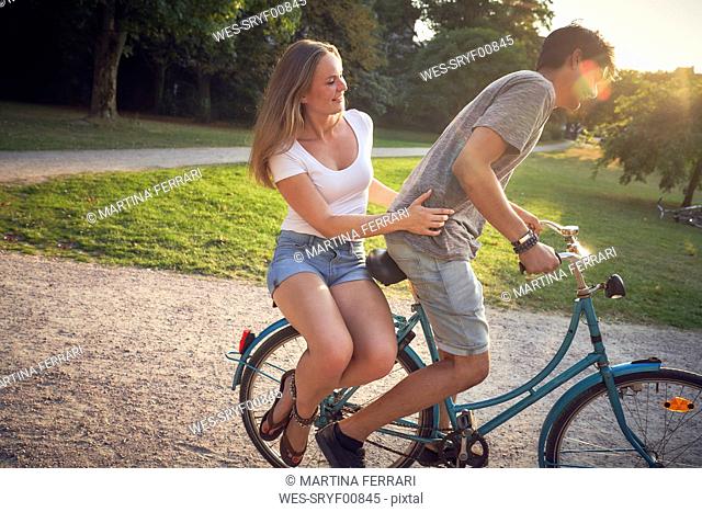 Young couple riding bicycle in park, woman sitting on rack