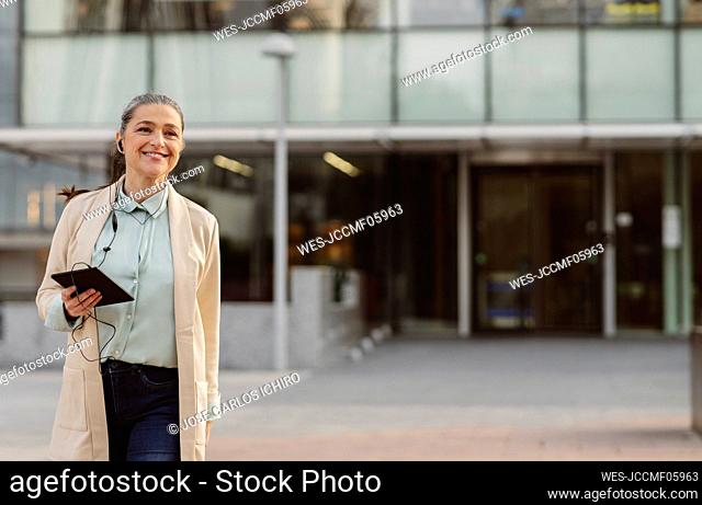 Happy businesswoman holding tablet PC listening music through in-ear headphones at office park