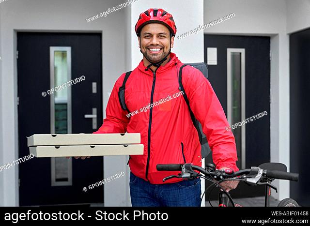 Smiling deliveryman with backpack holding pizza boxes and bicycle in front of house