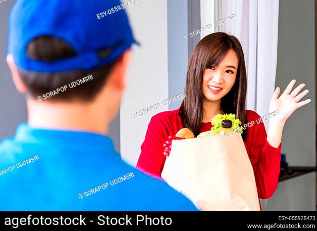 Asian young delivery man in blue uniform making grocery service giving fresh vegetables and fruits and food in paper bag to woman customer at front house after...